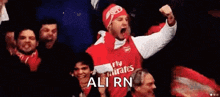 a man in a red fly emirates jersey is holding his fist in the air while sitting in a crowd of people .