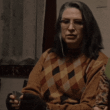 a woman wearing glasses and a plaid sweater holds a cigarette in her hand