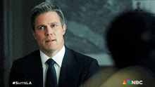 a man in a suit and tie is talking to another man with the nbc logo behind him