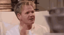a man in a white shirt is making a funny face while sitting in a chair in a kitchen .