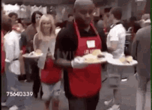 a man in a red apron is carrying plates of food in a crowd .