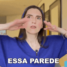 a woman in a blue sweater says " essa parede " with her hands on her face