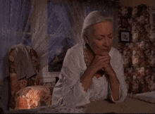 an elderly woman is sitting at a table with her hands folded in prayer