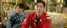 two young men are sitting on a couch with confetti falling around them . one of the men is wearing a red jacket .