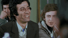 a man in a suit is smiling while sitting in a room with other men