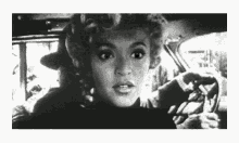a black and white photo of a woman in a car with a man in the background .