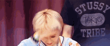a young man with blonde hair is wearing a stussy tokyo tribe shirt and a necklace .