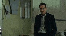 a man in a suit stands in a room with a white car in the background