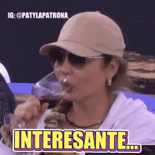 a woman wearing a hat and sunglasses drinking a glass of wine with the words interesante written on the bottom