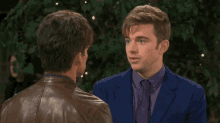 a man in a blue suit and purple tie talks to another man in a leather jacket