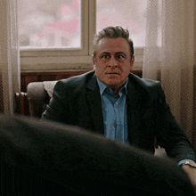a man in a suit and blue shirt sits in a chair in front of a window