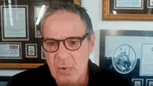 a man wearing glasses and a black shirt is standing in front of a wall with framed pictures on it .