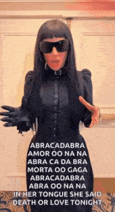 a woman wearing sunglasses and a black dress is standing in front of a wall .