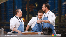 three men in aprons are standing around a counter with the nbc logo on the bottom right