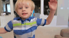 a young boy wearing a blue and white striped shirt is waving his hand in a living room .