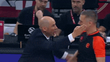 a man in a suit and tie is talking to another man in a red and black shirt .