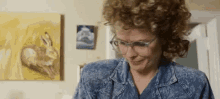 a woman with curly hair and glasses is smiling in front of a painting of a rabbit on the wall .