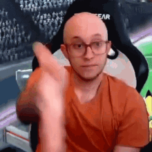 a bald man wearing glasses and an orange shirt is sitting in a chair and giving a thumbs up .