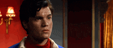 a young man in a blue jacket and red turtleneck looks at the camera