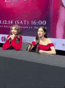 two women sitting at a table in front of a sign that says 12.14 sat 16:00