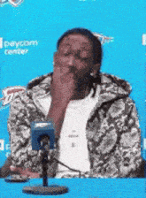 a man is sitting at a table in front of a microphone and covering his mouth with his hand .