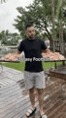 a man in a black shirt and grey shorts is standing on a deck holding a large piece of meat .