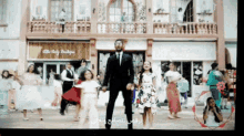 a man in a suit and tie stands in front of a group of children dancing