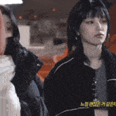 a woman in a black jacket stands next to another woman with korean writing on the bottom