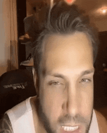a man with a beard wearing a white tank top is smiling and looking at the camera .