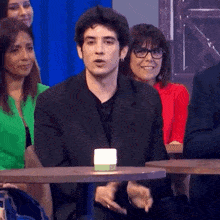 a man in a suit and glasses is sitting at a table in front of a crowd of people .