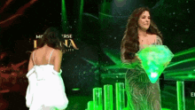 a woman in a white dress stands next to a woman in a green dress holding a diamond