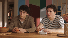 two young men sitting at a table with a bowl of fruit