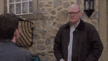 a bald man wearing glasses stands in front of a stone wall