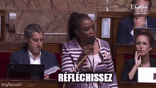 a woman is speaking into a microphone in a parliament while sitting in a row of people .