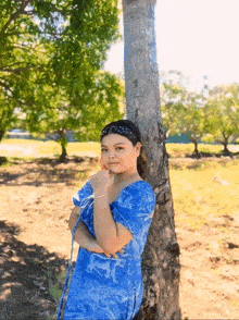 a girl in a blue dress leaning against a tree