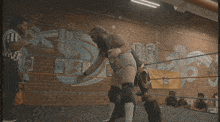 a referee watches two women wrestle in a ring with a mural on the wall behind them with the number 25 on it
