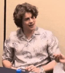 a young man is sitting at a table wearing a hawaiian shirt and a watch .