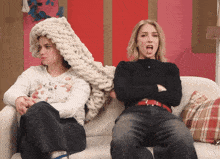 two women are sitting on a couch with their arms crossed one has a knitted hat on her head