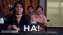 a woman is sitting at a desk in a classroom while a group of people are sitting in the background .