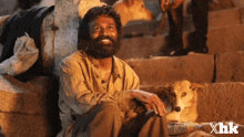 a man with a beard is sitting next to a dog with xhk written on the bottom right