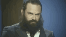 a man with a beard and long hair is wearing a suit and tie and looking at the camera .