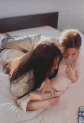 two women are laying on a bed reading a book