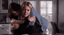 two women are hugging each other in a kitchen in front of a window .