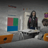 a girl in a leather jacket stands in a classroom with a boy sitting at a desk