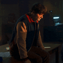 a man in a blue and red jacket sits at a table in a dark room