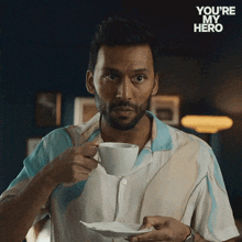 a man is drinking a cup of coffee with the words " you 're my hero " behind him