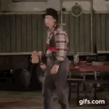 a woman in a plaid shirt and overalls is dancing in front of a table and chairs .