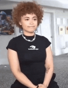 a woman with curly hair is wearing a black shirt and a necklace while sitting on the floor .