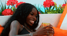 a woman in a black top is smiling while holding a drink in front of red flowers