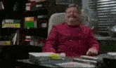 a man in a red shirt is sitting at a desk in an office with a mustache .